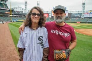 Josue and his father standing on a baseball field. Josue is a young man with shoulder-length brown hair and he is wearing a Chicago White Sox jersey. His dad puts his arm around Josue's shoulders and is wearing a red UChicago shirt. The White Sox stadium is visible in the background.