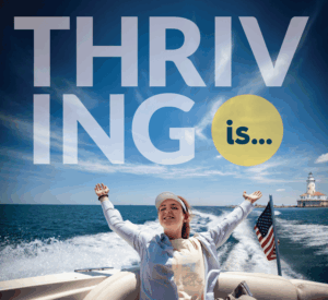 A young woman is on the stern of a boat with her arms fully extended as the wind blows in her hair and the sun shines on her face. Text reads: Thriving Is...