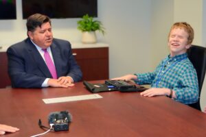 Governor J.B. Pritzker being interviewed by a young man an a plaid shirt who appears to be blind. Audio recording equipment sits on the table between them.