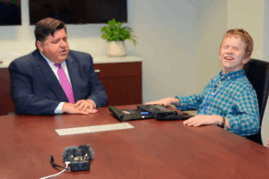Governor J.B. Pritzker being interviewed by a young man an a plaid shirt who appears to be blind. Audio recording equipment sits on the table between them.