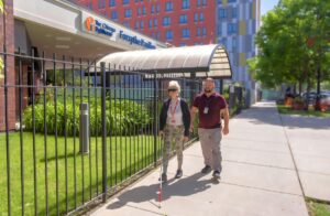 Two individuals walking outside The Chicago Lighthouse entrance, with one person using a white cane. They are surrounded by lush greenery and modern buildings on a sunny day.
