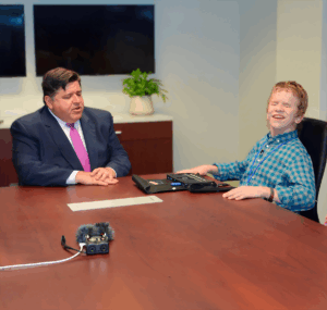 Governor J.B. Pritzker being interviewed by a young man an a plaid shirt who appears to be blind. Audio recording equipment sits on the table between them.
