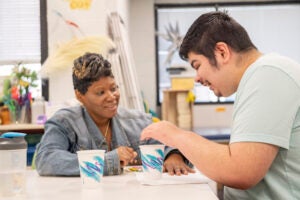 Sitting at a desk, a woman watches a visually impaired young man reach for a cup.