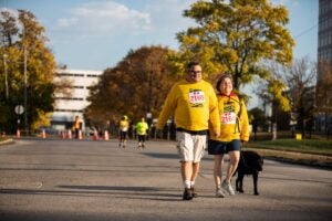 A man, woman, and dog walk outside. They are wearing yellow "Rise N Shine" race shirts.