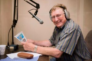 Person sitting at a broadcast desk with a microphone, wearing headphones, and holding a postcard, smiling. There's a mug and papers on the desk.