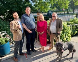 Greg and Turner stand with Lighthouse president and CEO Dr. Janet Szlyk, Ray C, and Peter T, who is holding a white cane. They are outside near a fence and greenery on a sunny day.