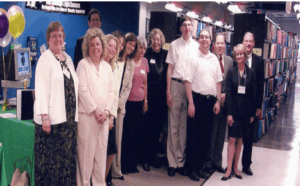 A group portrait from a news article (circa 2003)) of a group of people in front of a display and the bookshelves in the Illinois Instructional Material Center.