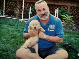 A photo of a man with short hair and a short, grey beard is sitting in a grassy area holding a fluffy beige puppy.