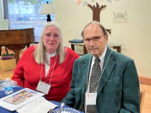 A man and a woman (Ken and Edith H.) are seated at a table, smiling towards the camera in a room with a piano and wall decorations.