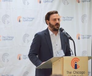 Peter speaking at a podium with The Chicago Lighthouse logos in the background. He has short brown hair and a short beard.