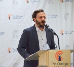 Peter speaking at a podium with The Chicago Lighthouse logos in the background. He has short brown hair and a short beard.