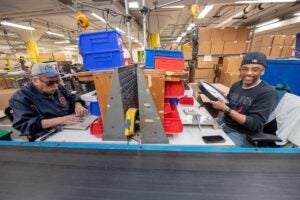 Two Lighthouse Industries employees are making clocks on a manufacturing assembly line. The man on the left is focused on assembling parts, while the man on the right is holding a product and smiling towards the camera.
