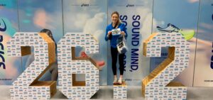 A young woman stands on a podium, holding a marathon bib and smiling.