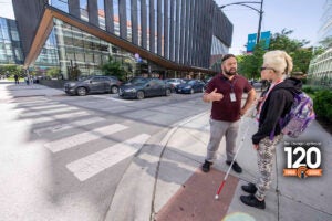 An Orientation & Mobility instructor teaches a visually impaired client how to navigate a busy intersection using a white cane.