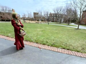 A young woman walking along a paved path on a college campus, holding a white cane. In the background, there are grassy areas and college buildings.