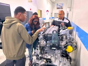 A group in a workshop examining a large engine. One person points to a part of the engine, while the two students holding white canes and wearing protective glasses experience the parts of the engine through touch.