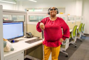 A woman standing confidently in an office cubicle, holding a white cane. She is wearing a headset and sunglasses, and computers and phones are visible in the background.