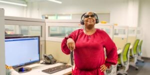 A woman standing confidently in an office cubicle, holding a white cane. She is wearing a headset and sunglasses, and computers and phones are visible in the background.