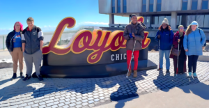 A group of six students and Lighthouse staff stand in front of a large "Loyola Chicago" sign with Lake Michigan and a modern building is visible in the background. Several people are holding white canes.