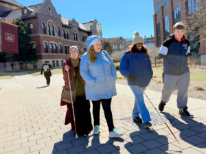 A group of four people with canes walks along a sunny brick path at Loyola University. A large building with a Loyola University Chicago banner is visible in the background. 