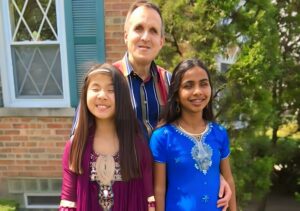 The Sprecher Family poses for a photo. The two daughters are wearing brightly colored dresses.