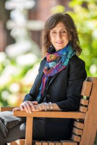 Julie Stark, sitting on a wooden bench, wearing a colorful scarf and dark clothing, against a blurred outdoor background. She is a white woman with shoulder-length brown hair and a kind smile.