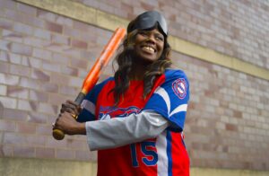 Kalari G. standing in front of a brick wall holding a baseball bat, wearing a jersey with the word "Comets" and the number 15. She has a black eye mask on her head and is smiling.