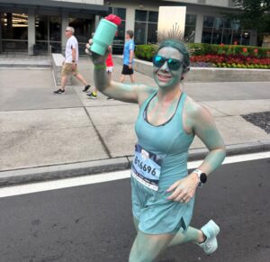 Darcie is painted blue, running a marathon, holding up her water bottle and smiling at the camera triumphantly.
