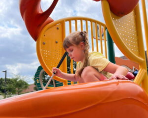 A blind child goes down a playground slide holding her white cane